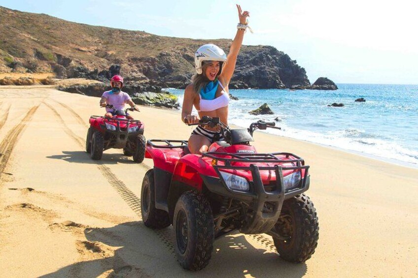 Two people riding red ATVs on a sandy beach with rocky cliffs and the ocean in the background. 2