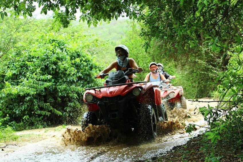 Two people riding an ATV through a muddy path surrounded by greenery. 4