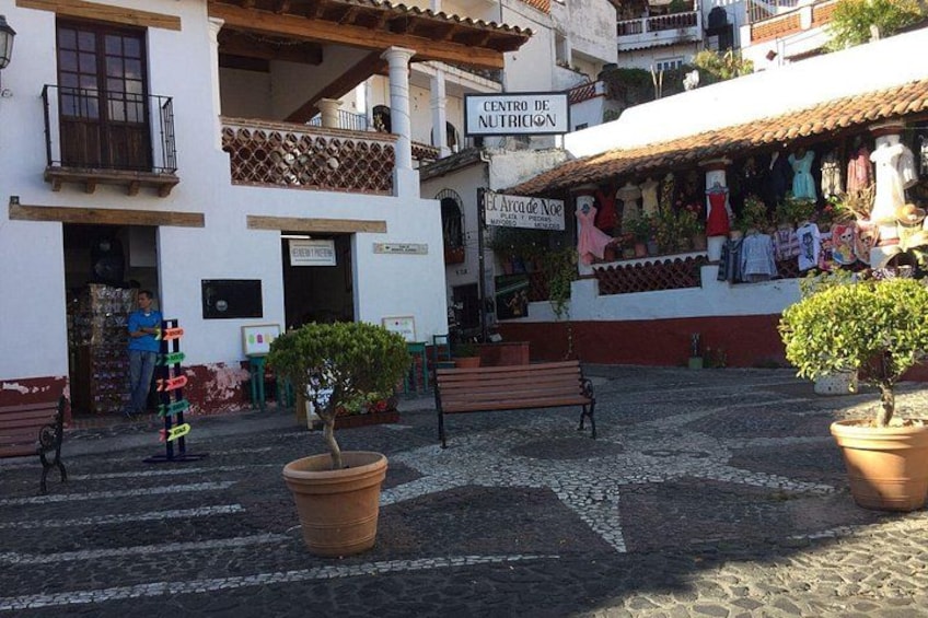 A cobblestone street with a sign for 'Centro de Nutricion,' a building with a balcony, and a shop with clothes displayed outside. 1