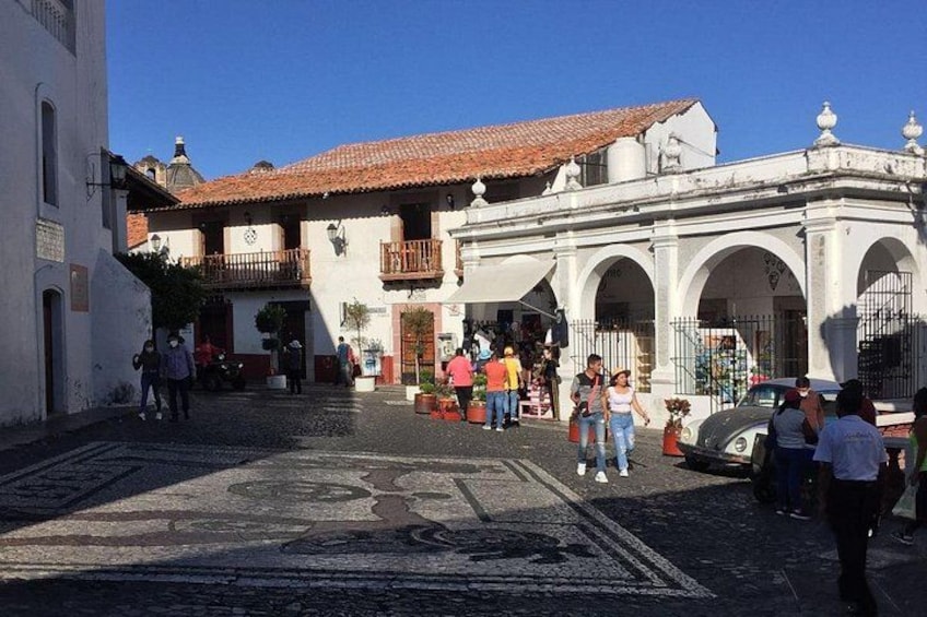A cobblestone street with pedestrians, a white building with a red-tiled roof, and a white structure with arches. 3
