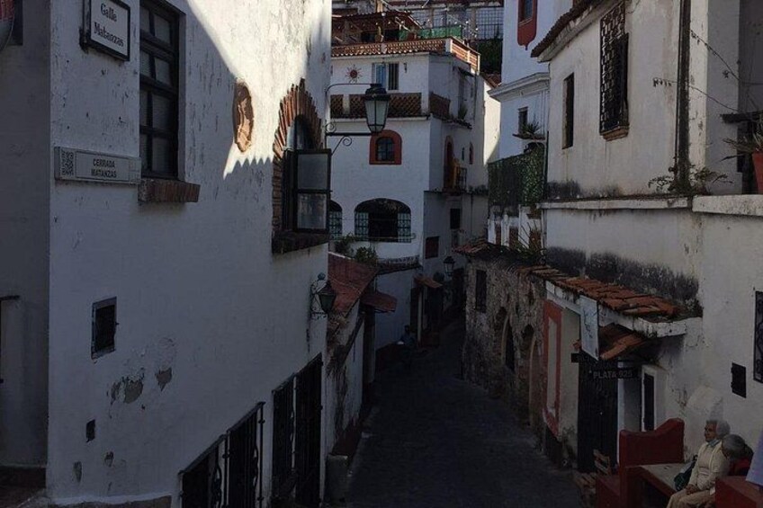 A narrow street flanked by white buildings with red-tiled roofs, narrow passageways, and a few visible signs. 4