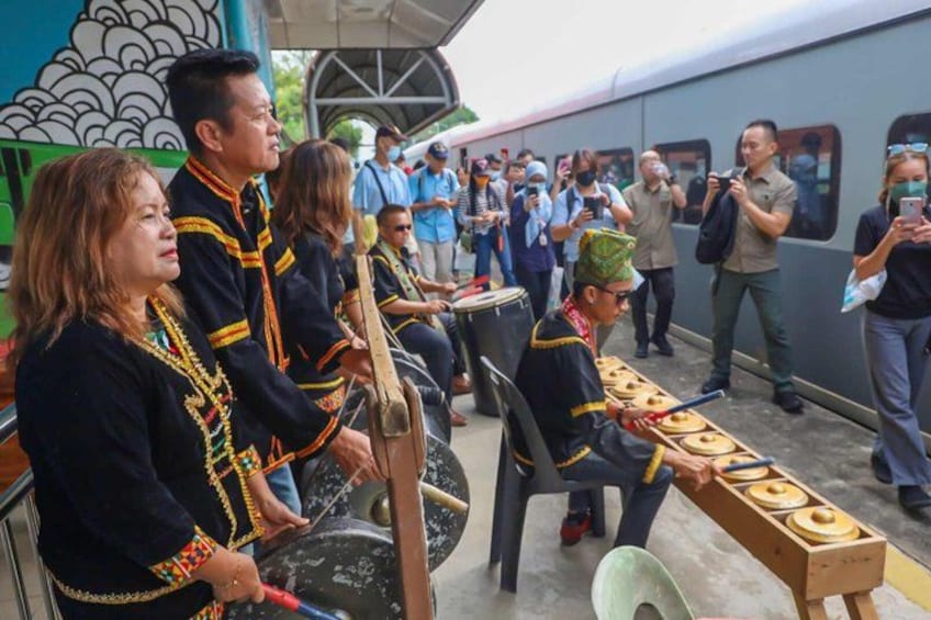 A group of people playing traditional instruments, seated on chairs, with a train in the background. 1