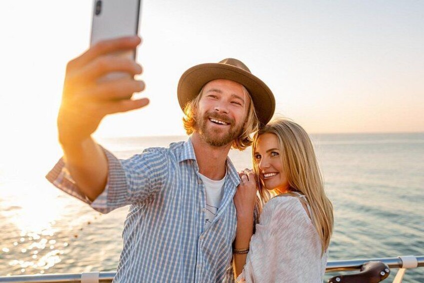 A person taking a selfie with a smartphone, wearing a hat, and a striped shirt, with a body of water and a clear sky in the background. 5