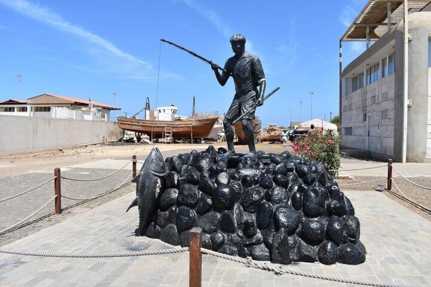 A statue of a figure with a spear, standing on a pile of fish, with a boat and buildings in the background. 4