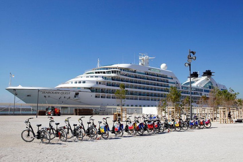 Et stort krydstogtskib, 'Sea Orient Quest', ligger til kaj ved en havn, hvor cykler står parkeret foran. 3
