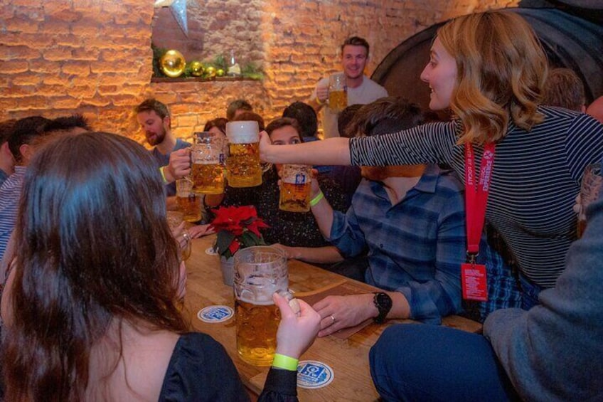 A group of people toasting with beer mugs at a wooden table in a rustic setting. 5
