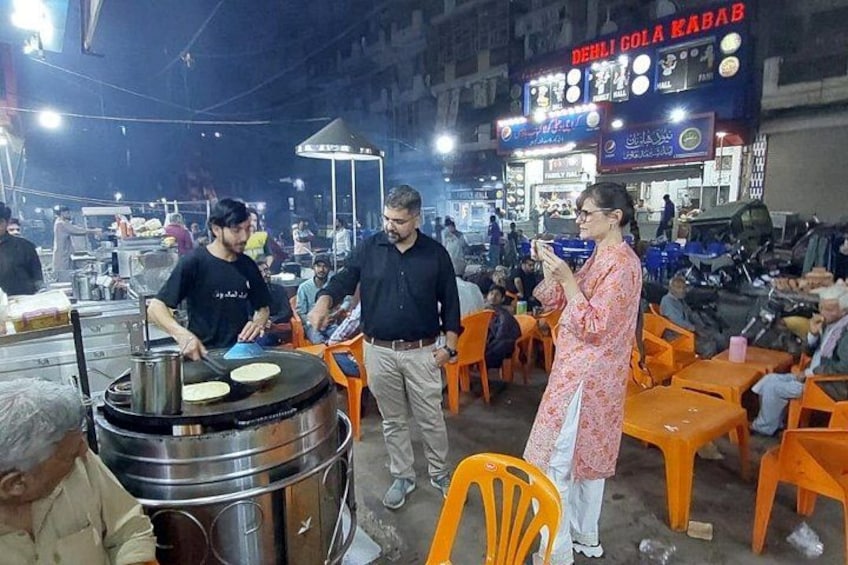 A street food vendor cooking on a large grill, a woman in traditional attire, and a busy street with orange chairs and tables. 5