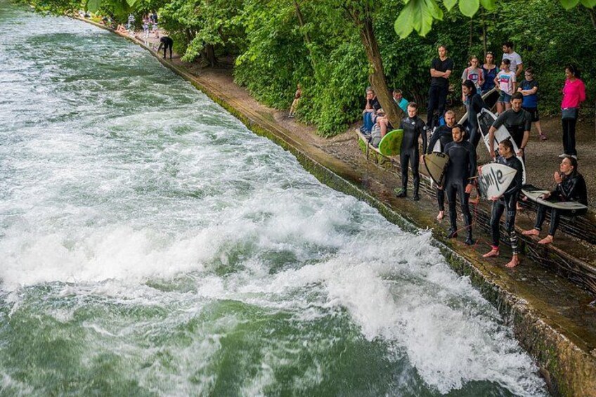 Een rivier met krachtige stromingen, een groep mensen in neopreenpakken met surfplanken, en iemand in een zwart neopreenpak die langs de oever loopt. 4