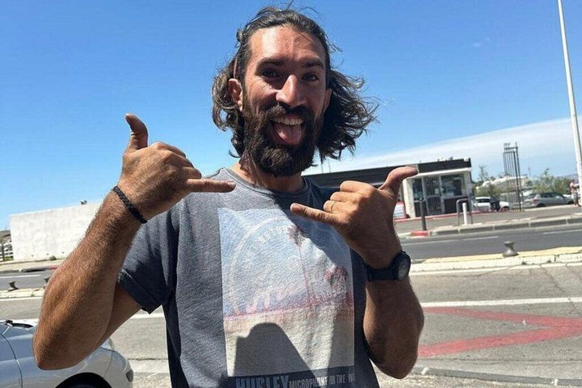 En person peger på sig selv med begge hænder, iført en T-shirt med et grafisk design, stående i et udendørs miljø med en klar himmel og en bygning i baggrunden. 3