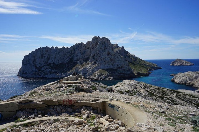A rocky coastline with a prominent limestone formation, a clear blue sea, and a small island in the distance. 2