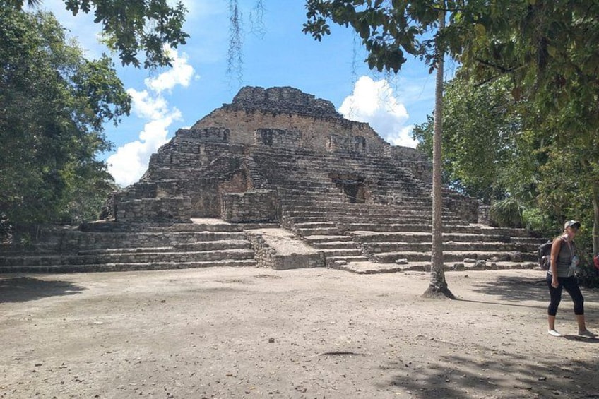 Une vaste structure ancienne, en forme de pyramide, avec un escalier menant à son sommet, entourée d’arbres et sous un ciel dégagé. 1