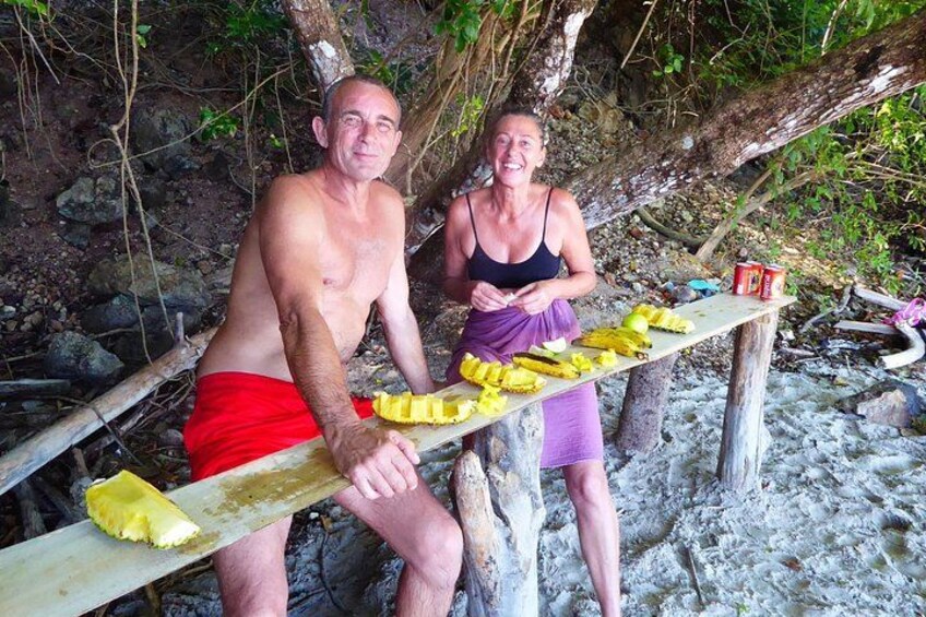 Two people sitting on a wooden bench outdoors, with a table in front of them holding food items. 2