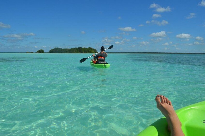 Iemand kajakt op helder turquoise water, met een groene opblaasbare kajak. Achtergrond: een andere kajak en een eiland in de verte. 1
