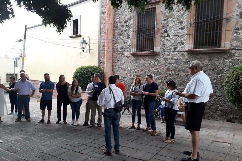 A group of people standing on a paved street, some engaged in conversation, with stone buildings and a lamp post in the background. 4