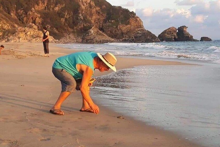 En person i blå skjorta och hatt som böjer sig framåt på en sandstrand med klippiga branter i bakgrunden. 5