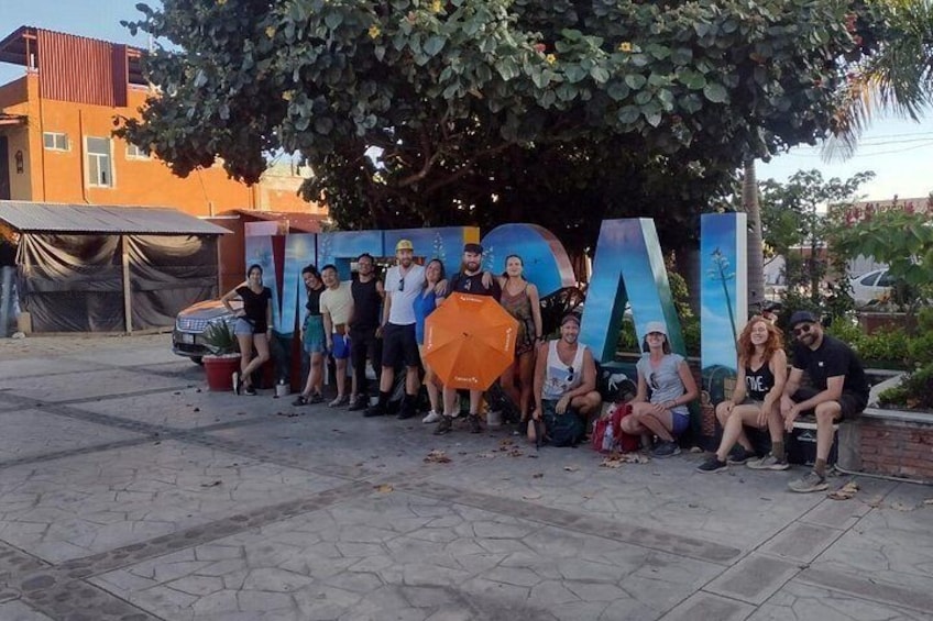 Eine Gruppe von Menschen posiert vor großen Buchstaben, die 'NATURAL' buchstabieren, mit einem Baum und Gebäuden im Hintergrund. 4