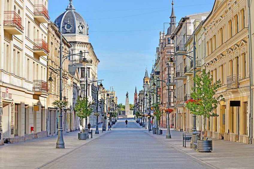 Eine breite Straße gesäumt von historischen Gebäuden, ein klarer Himmel und Bäume mit roten Blüten. 1