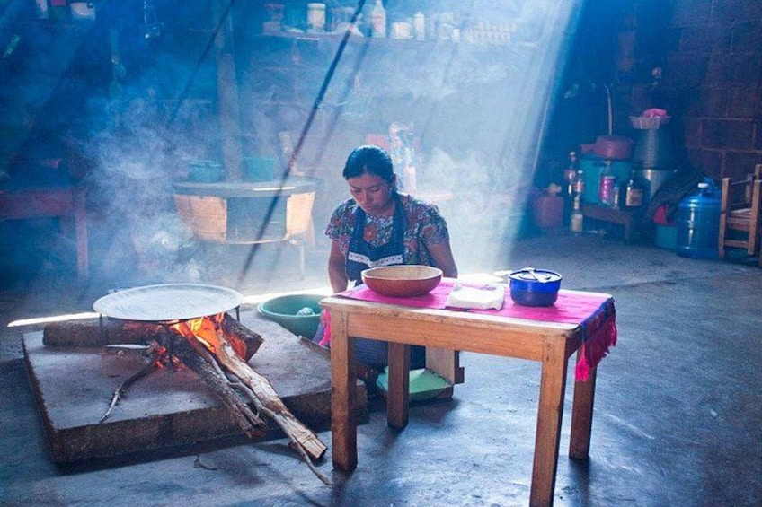 Une personne cuisine au-dessus d’un feu de bois dans une cuisine rustique, avec une table, des casseroles et un grand pot en arrière-plan. 1