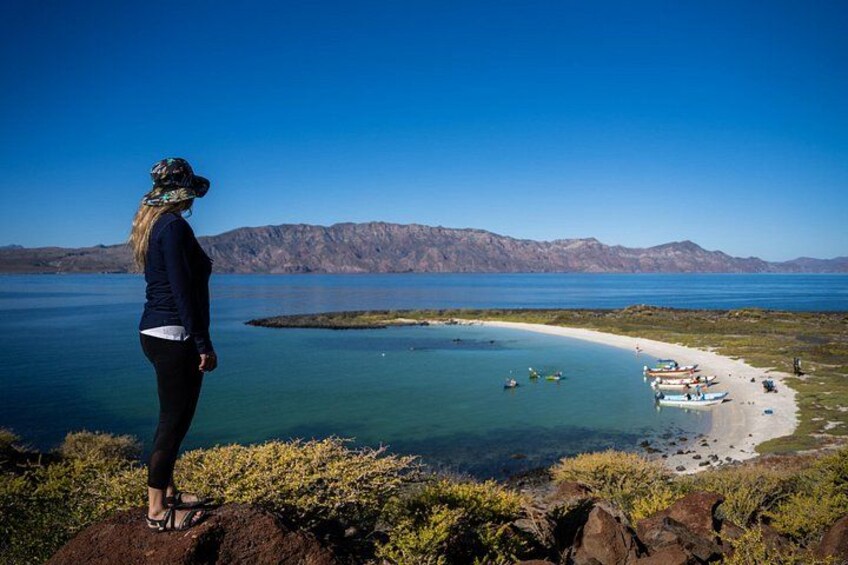 En person står på en klippavsats och blickar ut över en strand med båtar och en klar, blå himmel. 3