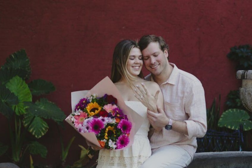 Una coppia che tiene un bouquet di fiori, seduta di fronte a un muro rosso con vegetazione sullo sfondo. 1