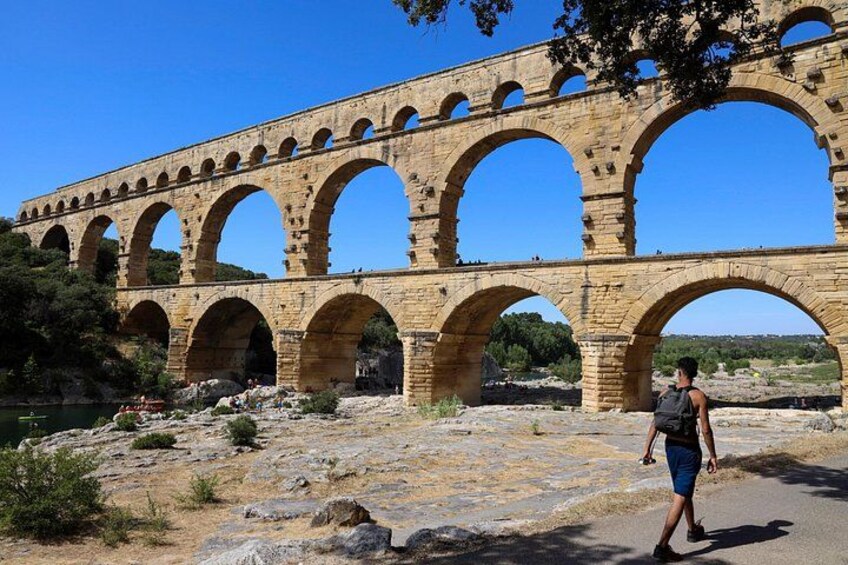 A stone aqueduct with multiple arches, a person walking on a path, and a clear blue sky. 1