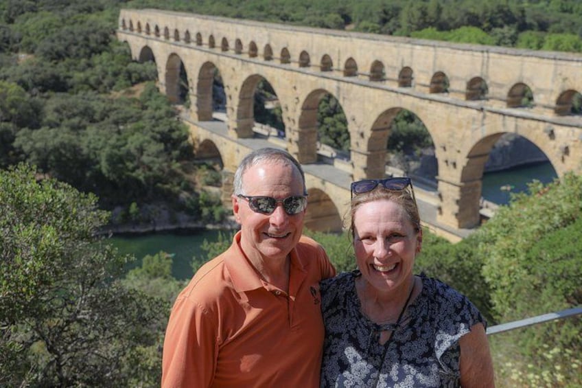 A man and a woman standing in front of a stone bridge with multiple arches over a river, surrounded by greenery. 3