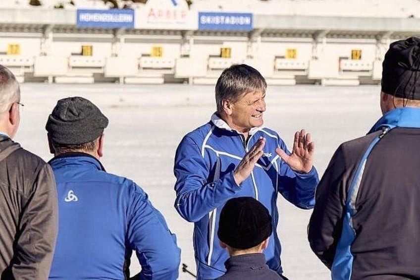 Una persona con una chaqueta azul gesticula, con un área de asientos de estadio al fondo. 1