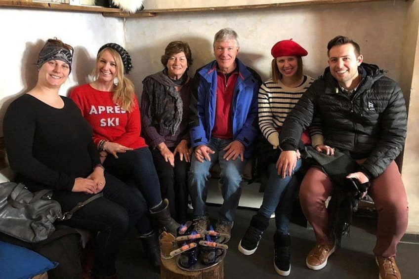 A group of people sitting on a bench in a room with a concrete wall and a wooden table with items on it. 2