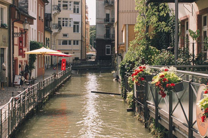 Ein schmaler Kanal, begrenzt von Gebäuden, eine Brücke mit Blumendekorationen und ein Café mit Sonnenschirm. 3