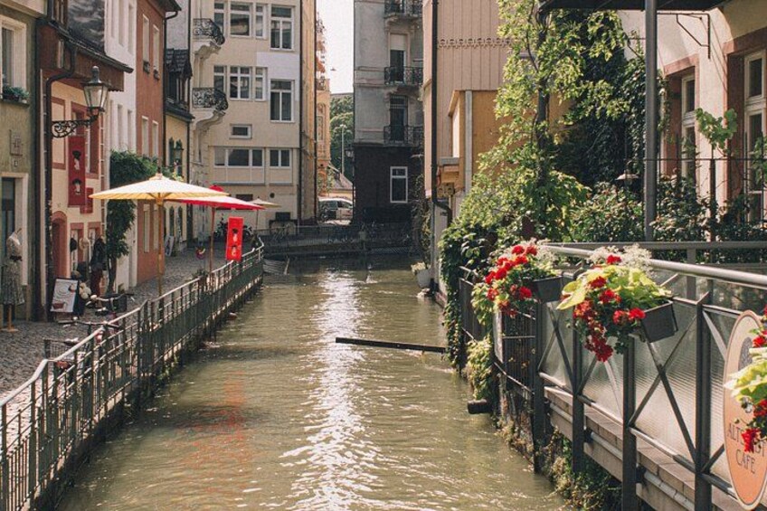 Un estrecho canal bordeado por edificios, un puente con barandillas y mesas al aire libre con sombrillas. 3