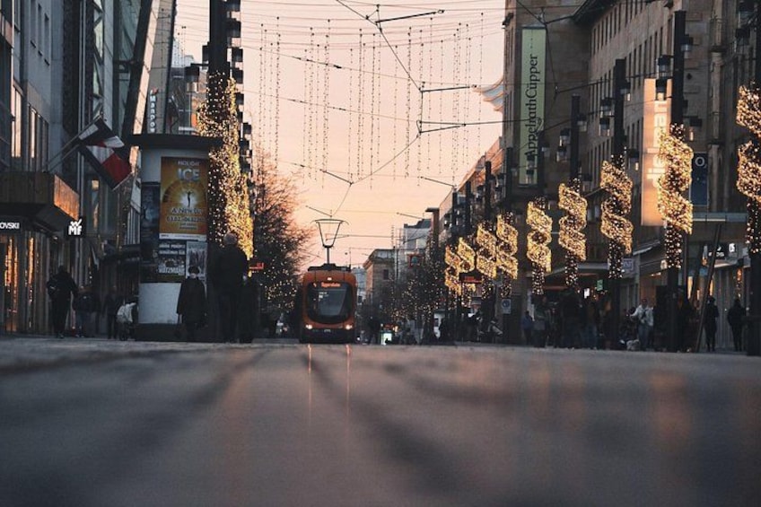A tram moving down a street lined with buildings and trees decorated with lights. 2