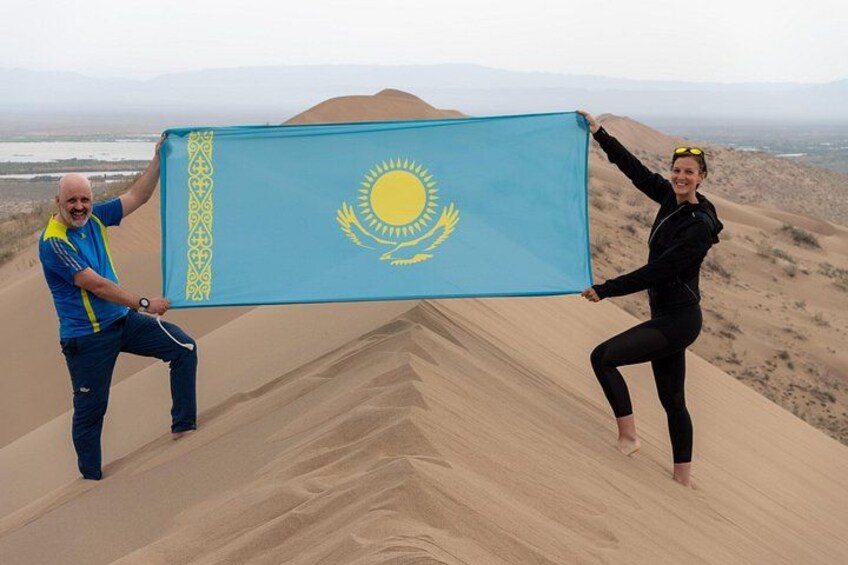 Twee personen houden een vlag vast op een zandduin, met een woestijnlandschap op de achtergrond. 2