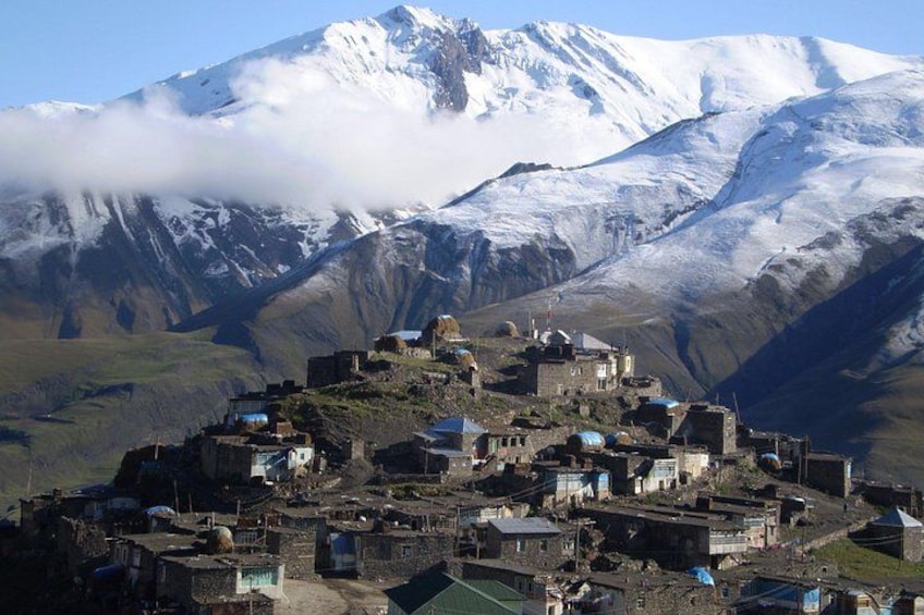 Un village de montagne avec des bâtiments construits sur le flanc d’une colline, un sommet enneigé en arrière-plan et un ciel dégagé. 2