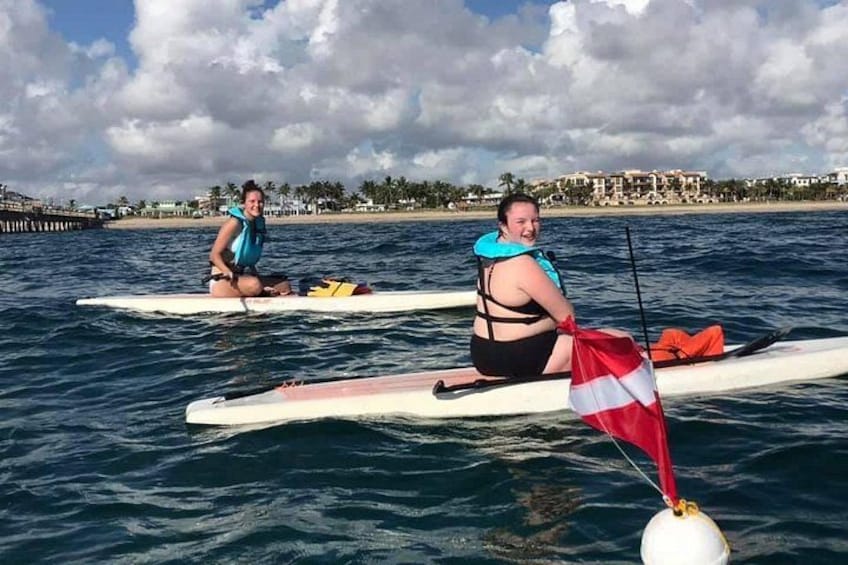 Dos personas en tablas de remo en aguas tranquilas, con una playa y edificios al fondo. 4