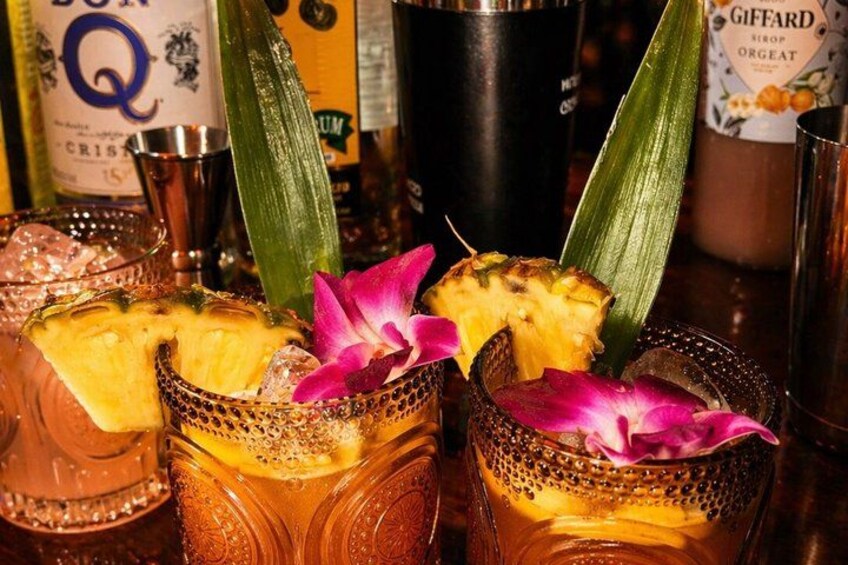 Tres cócteles con rodajas de piña y flores moradas, decorados con hojas verdes, en un bar con botellas al fondo. 2