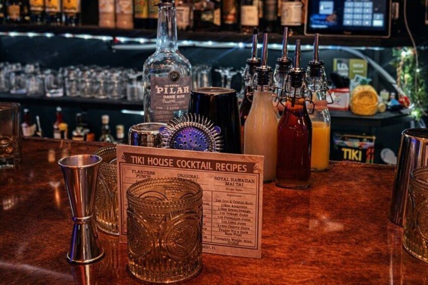 Un bar con un cartel de recetas de cócteles Tiki House, varias botellas y vasos. 4