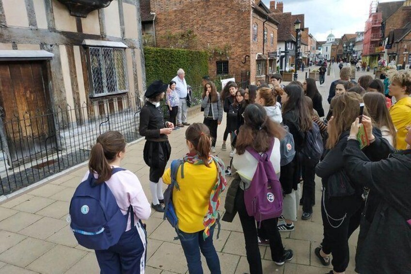 A group of people standing on a street, some with backpacks, in an urban area with buildings and a fence. 5