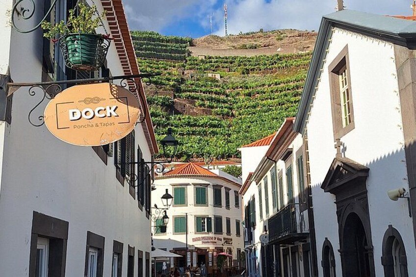 Une rue bordée d’immeubles, une enseigne indiquant « DOCK Poncha & Tapas », et des vignobles en arrière-plan. 2