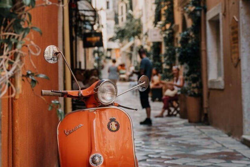 A vintage orange Vespa parked on a narrow street with people in the background. 1