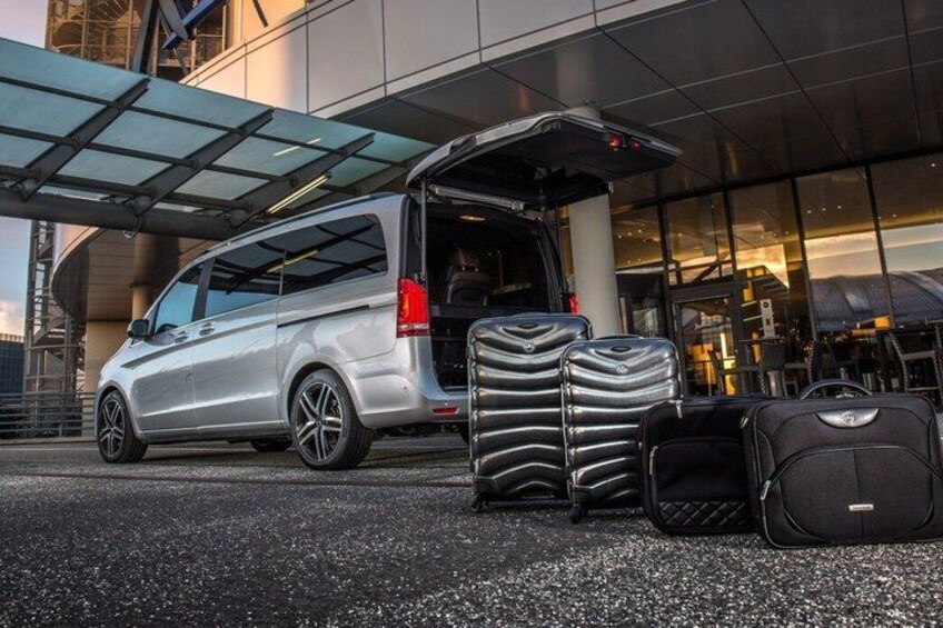 A silver minivan parked in front of a building with its rear door open, luggage stacked inside, and a clear sky in the background. 1