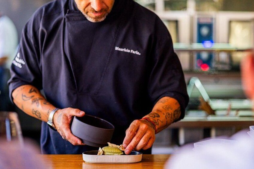 A person in a black apron with tattoos, holding a bowl and a plate with food. 5