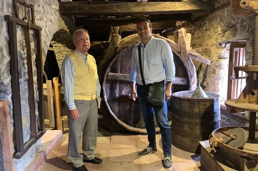 Two people standing in a rustic interior with stone walls, wooden barrels, and a large wooden wheel. 4