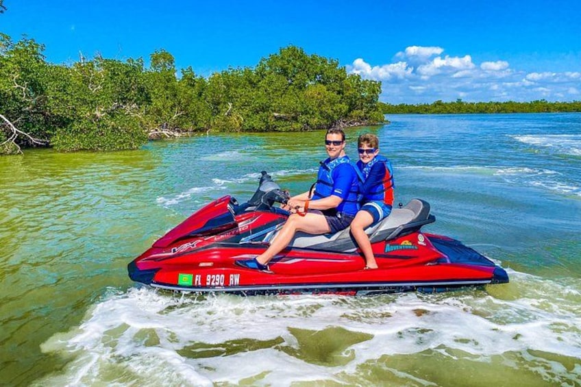 Zwei Personen auf einem roten Jetski im klaren Wasser, mit bewaldeter Küstenlinie. 4