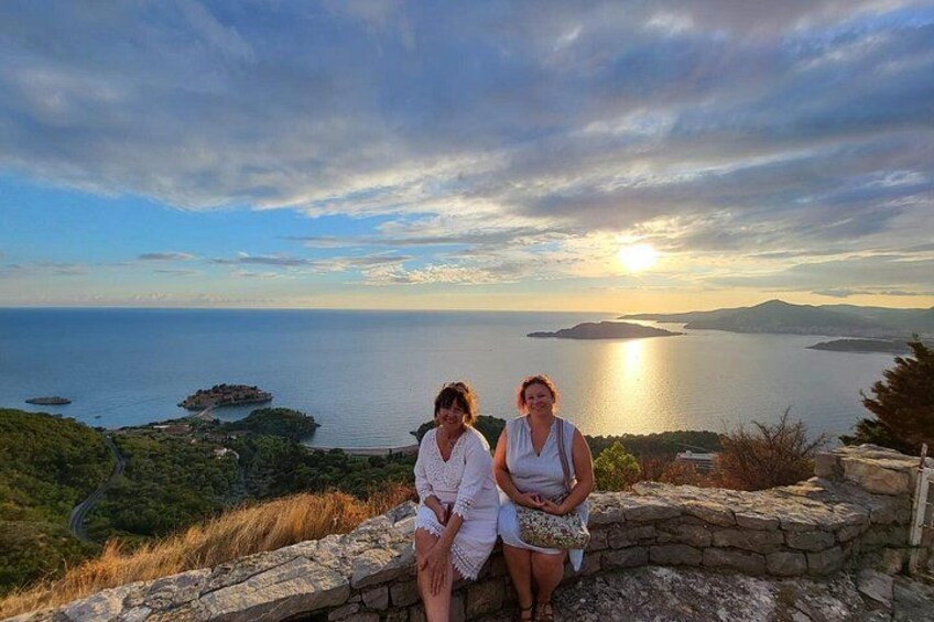 Two people sitting on a stone wall overlooking a coastal landscape with a sunset. 3