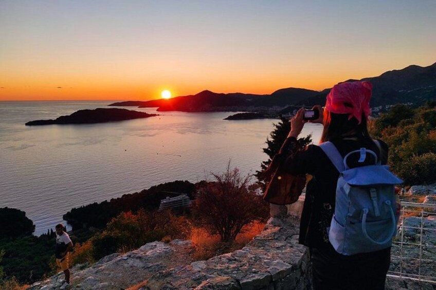A sunset over a body of water with islands, a person taking a photo, and another person walking. 2