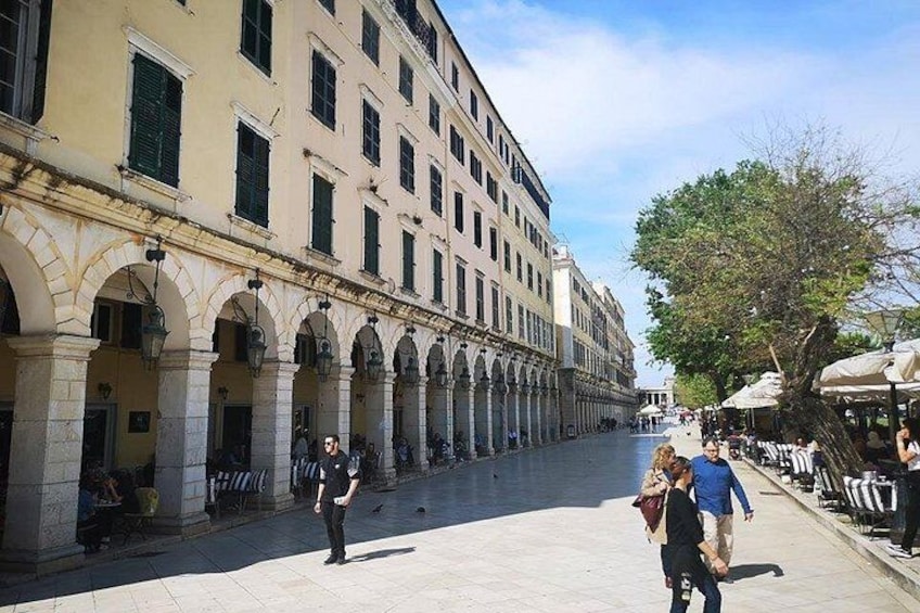 A wide pedestrian street with historic buildings, a tree-lined sidewalk, and outdoor seating. 1
