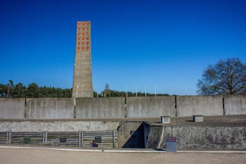 A tall concrete monument with red symbols, surrounded by a low wall and a paved area with benches. 1
