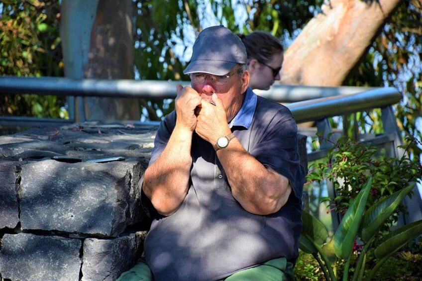 Un hombre sentado en un banco de piedra, con gorra y reloj, con follaje y una barandilla de metal al fondo. 4