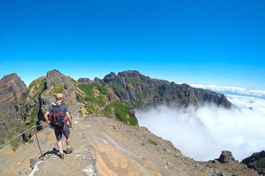 Ein Wanderer auf einem Bergpfad mit felsigem Gelände und Blick auf Wolken darunter. 3