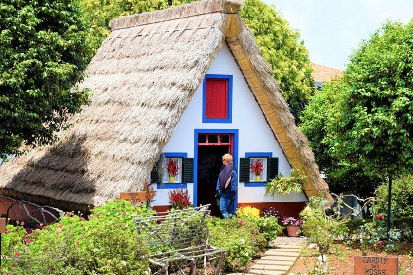 Une maison à toit de chaume, avec une porte bleue et des volets rouges, entourée d’une végétation luxuriante et d’un jardin. 3